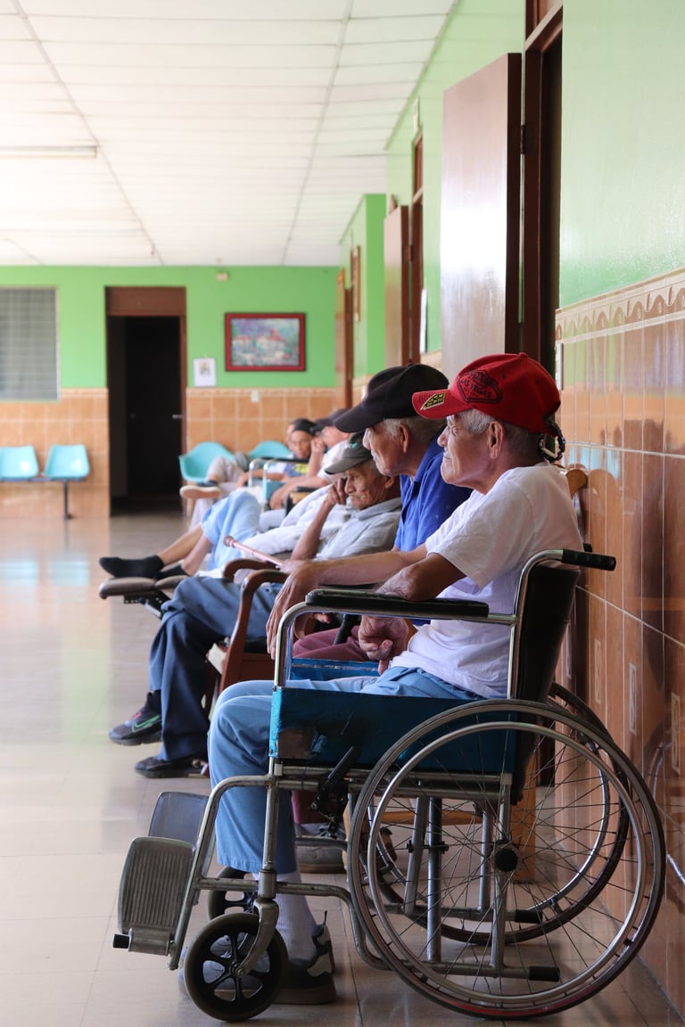 Pacientes adultos mayores en pasillo de hospital, con hombre en silla de ruedas usando gorra roja