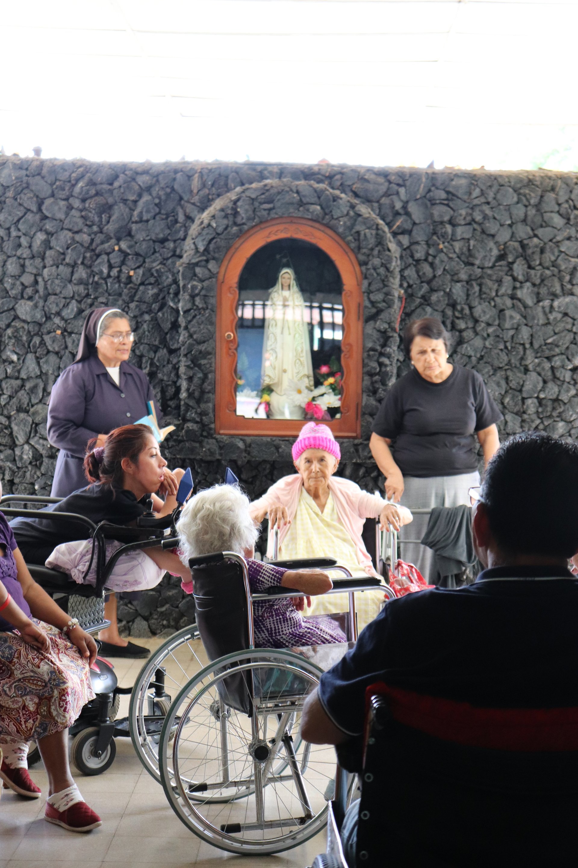 Grupo reunidos al aire libre en santuario religioso con ventana arqueada, incluyendo mujer adulta mayor en silla de ruedas y varios asistentes con ropa casual